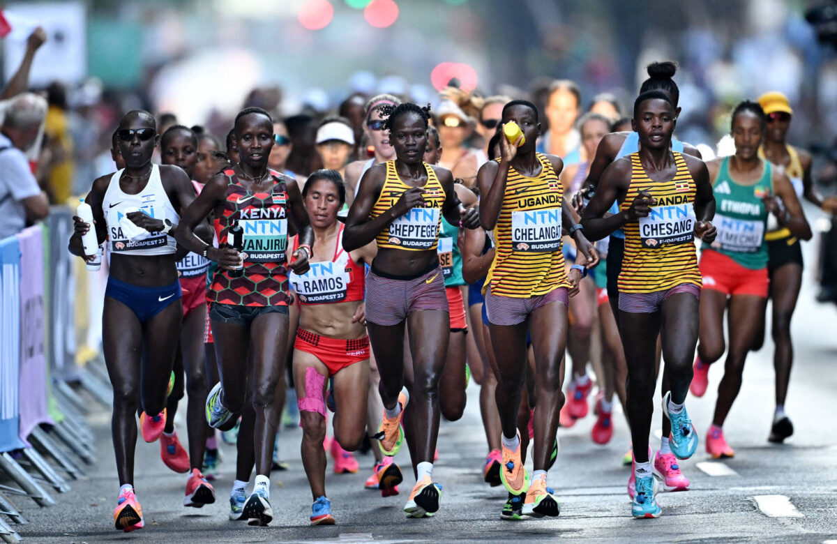 A imagem mostra um grupo de mulheres competindo em uma corrida. Elas estão vestidas com trajes de atletismo, com algumas usando uniformes de diferentes países, como Kenya e Uganda. O cenário é urbano, com espectadores ao fundo e uma atmosfera de competição. As corredoras estão em movimento, algumas com expressões de concentração.