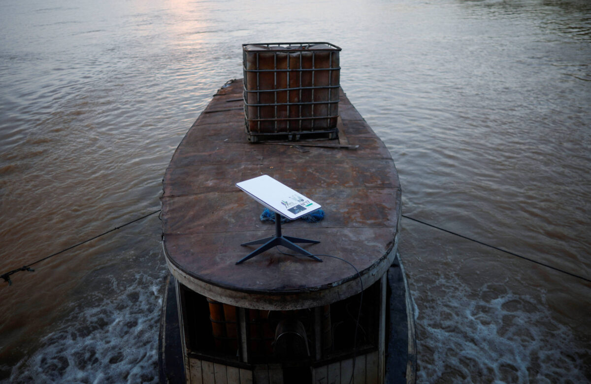 A imagem mostra a parte superior de um barco flutuando em água. No centro, há um equipamento da Starlink parecido com um tablet, a superfície é branca com pernas pretas, e ao fundo, uma cesta de material em cima do barco. A água ao redor reflete a luz do sol, criando um ambiente tranquilo.