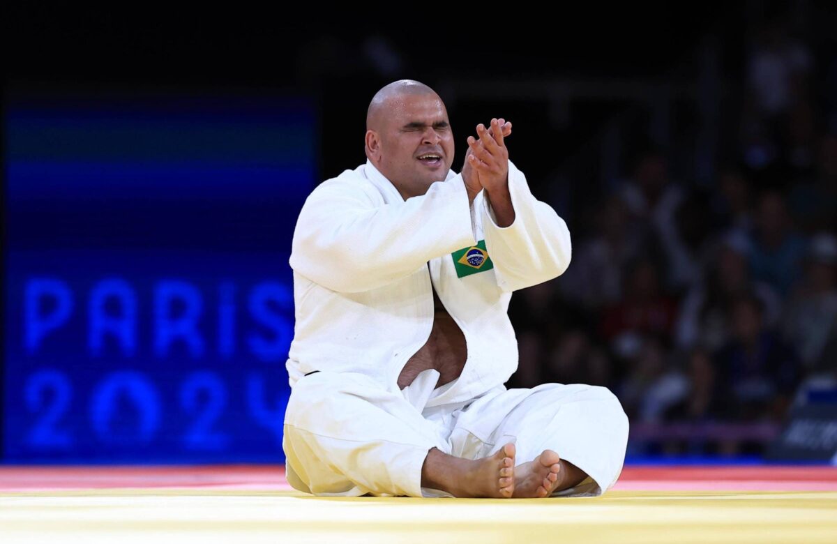 Um atleta de judô está sentado no tatame, aplaudindo. Ele usa um judogi branco e tem uma bandeira do Brasil em sua roupa. O fundo mostra uma tela com as palavras