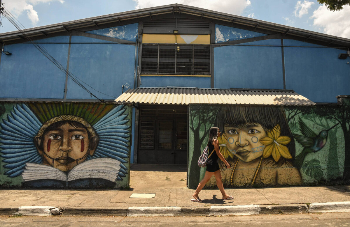 A imagem mostra a frente da escola, um edifício azul com um telhado de metal. Na parede à esquerda, há um mural que retrata um menino indígena com cocar e um livro, enquanto à direita há um mural de uma menina indígena com flores no cabelo. Uma jovem está caminhando na calçada em frente à escola, carregando uma mochila nas costas.