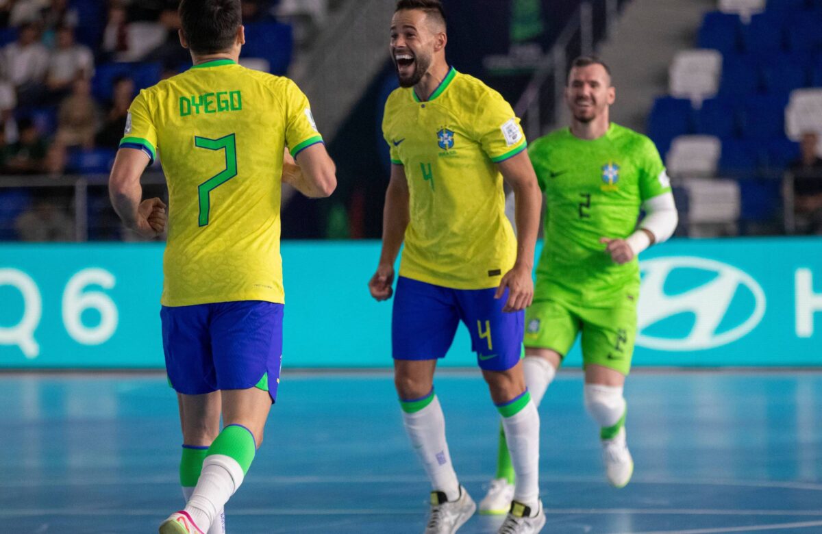 Três jogadores de futsal da seleção brasileira estão em um momento de celebração em quadra. O jogador à esquerda, com a camisa amarela e o número 7, é Dyego. O jogador central, também com a camisa amarela e o número 4, está sorrindo e levantando a mão. O terceiro jogador, à direita, usa uma camisa verde e parece estar se juntando à celebração. O fundo mostra uma quadra de futsal com uma iluminação suave e uma tela publicitária.