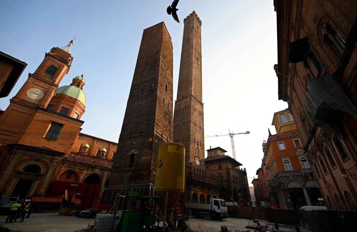 A imagem mostra as famosas Torres de Bolonha, duas torres inclinadas, em um ambiente urbano. Ao fundo, há uma cúpula de uma igreja e outros edifícios históricos. O céu está claro, indicando um dia ensolarado, e há um pássaro voando acima das torres.