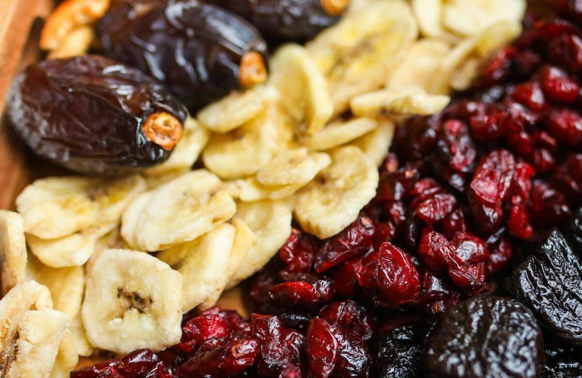 A imagem mostra uma variedade de frutas secas e nozes dispostas em uma bandeja de madeira. À esquerda, há tâmaras escuras e fatias de banana desidratada. No centro, estão cranberries vermelhas e secas. À direita, há ameixas secas escuras e nozes pecan marrons.