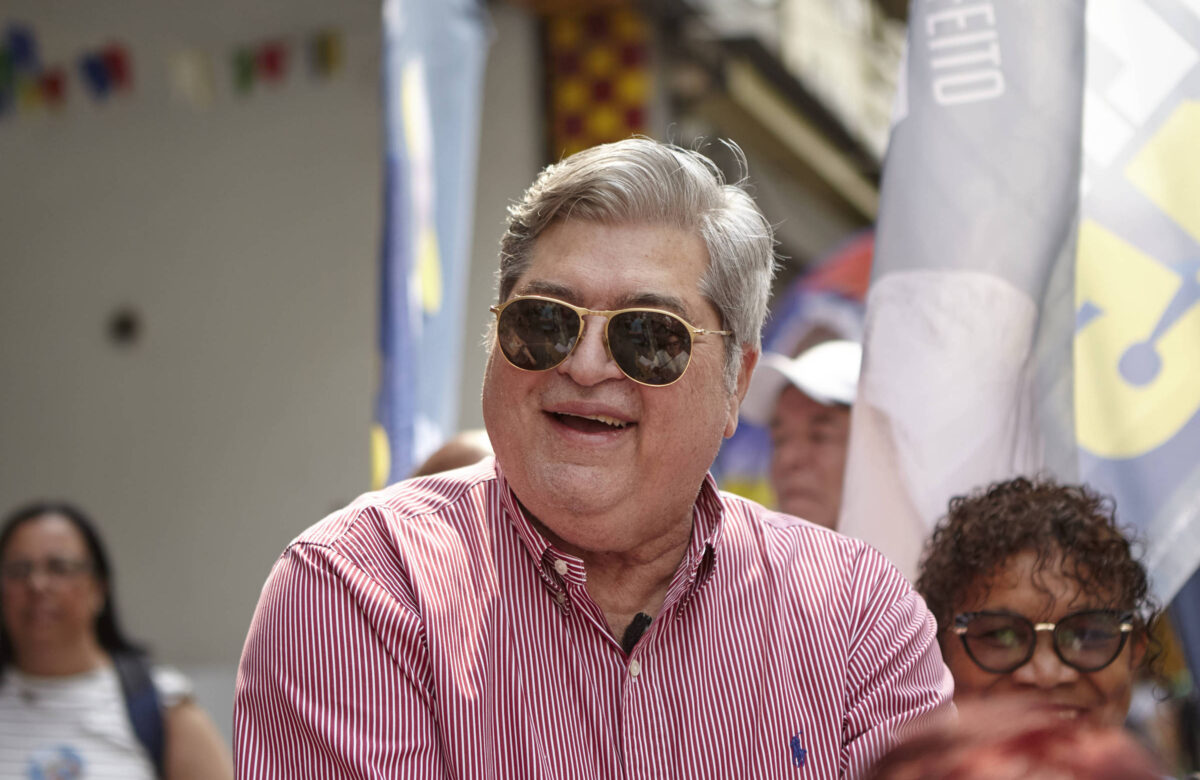 Datena, um homem de cabelos grisalhos, usando óculos escuros e uma camisa rosa, está sorrindo em um evento ao ar livre. Ao fundo, há pessoas e bandeiras visíveis, sugerindo um ambiente festivo ou de celebração.