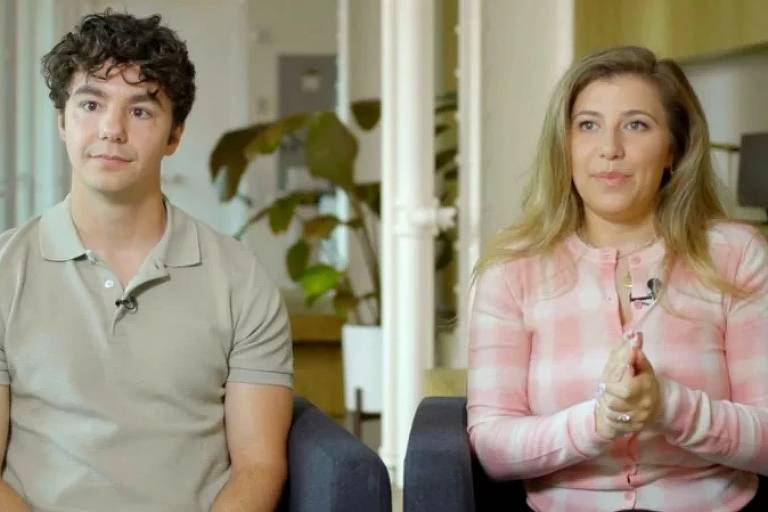 A imagem mostra duas pessoas sentadas em um ambiente moderno. À esquerda, um homem com cabelo cacheado e uma camiseta de cor clara, olhando para frente. À direita, uma mulher com cabelo longo e liso, vestindo um suéter rosa com listras claras, que está gesticulando com as mãos. Ao fundo, há plantas e uma mesa, sugerindo um espaço de trabalho ou uma sala de estar.