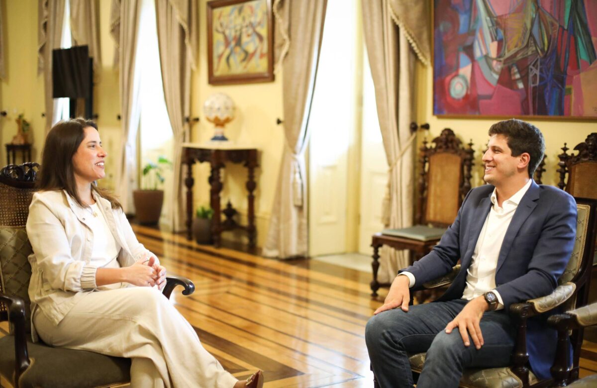 Raquel Lyra sentada vestida de branco olha para o prefeito João Campos, que também está em uma cadeira com terno azul e camisa branca. Ambos sorriem e estão em uma sala fechada