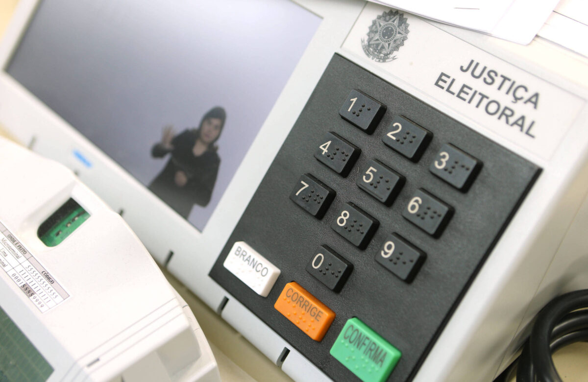 Close em uma urna eletrônica e em um aparelho de biometria. No canto da tela da urna, uma intérprete sinaliza em Libras