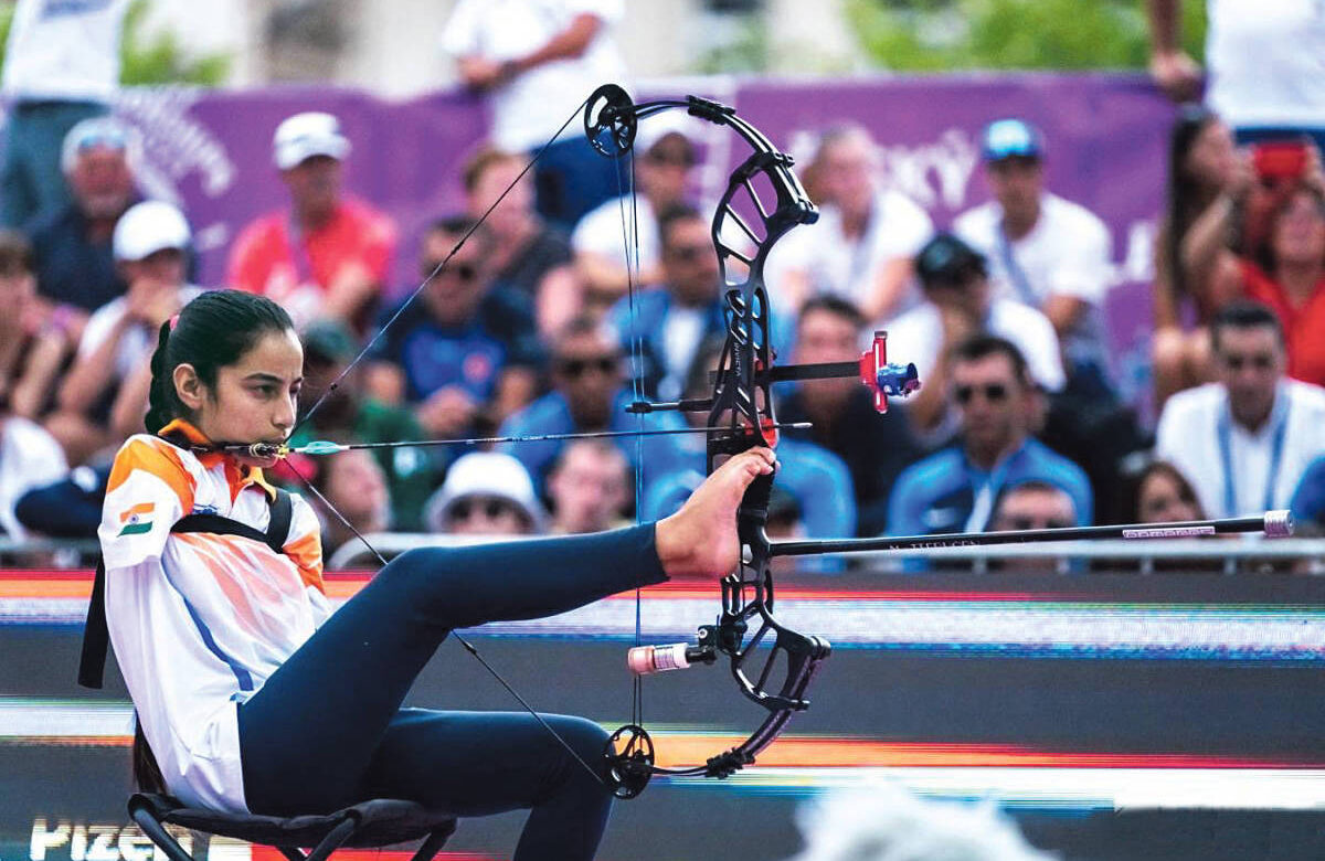 A imagem mostra uma arqueira em uma competição de tiro com arco, utilizando os pés para manusear o arco e flecha, ao invés das mãos. Ela está sentada em uma cadeira, usando uma camiseta com as cores e emblema da bandeira da Índia. Ao fundo, é possível ver várias pessoas assistindo à competição