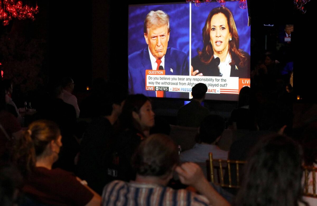 A imagem mostra um grupo de pessoas assistindo a um debate político em uma tela grande. Na tela, estão duas figuras, Trump e Kamala, discutindo. O ambiente é escuro, com algumas luzes ao fundo. As pessoas estão sentadas, algumas de costas para a câmera, e a atenção delas está voltada para a tela.