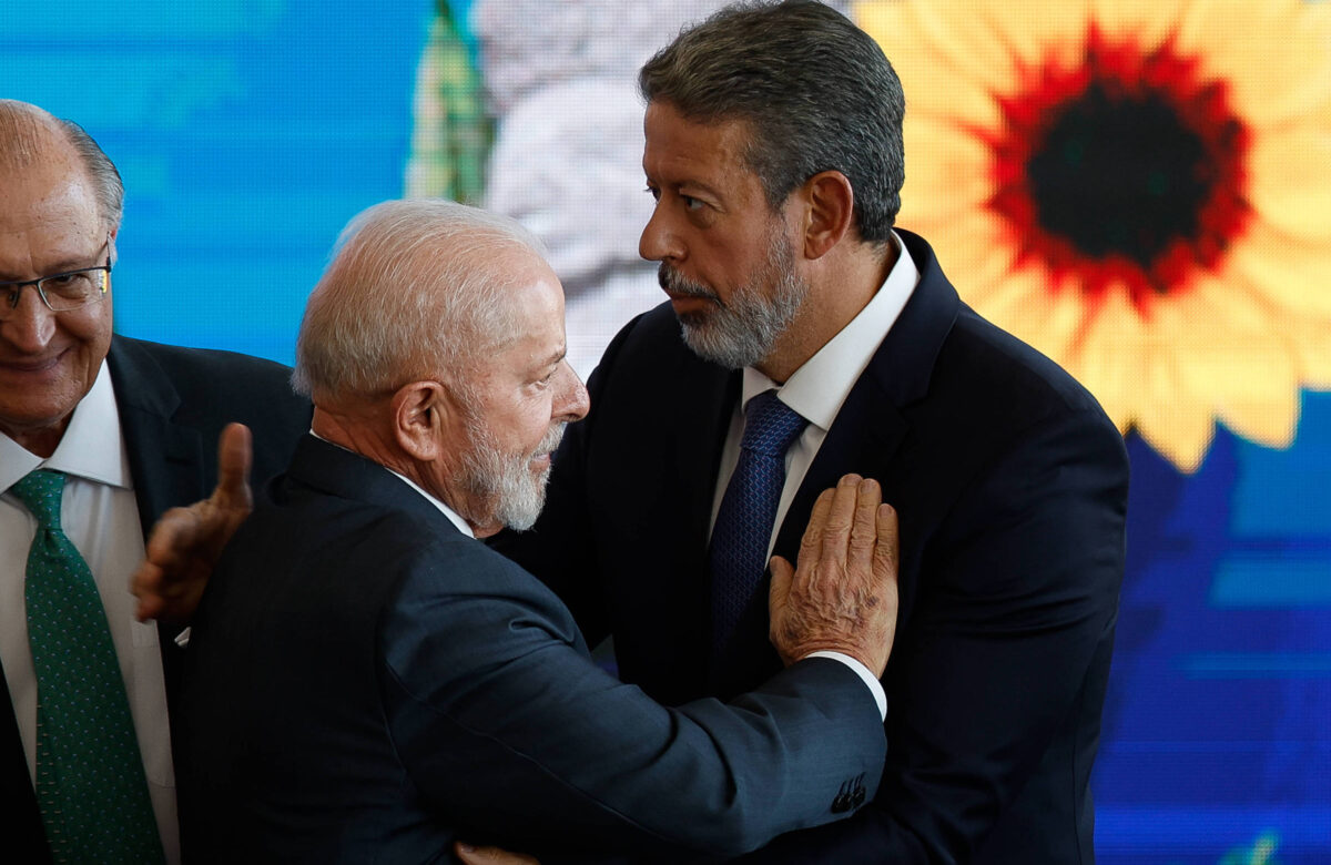 Lula, um homem branco de barba e cabelos ralos da mesma cor, coloca a mão sobre o peito de Lira, um homem branco, de barba e cabelo castanhos grisalhos. Os dois vestem terno e gravata escuros e ao fundo, ao lado deles, está o Alckmin, um homem branco, calvo, com terno e gravata verde. No telão, uma grande flor com pétalas amarelas