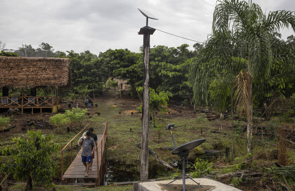 A imagem mostra uma cena rural com uma pessoa caminhando por uma ponte de madeira, enquanto ao fundo se observa uma casa simples e vegetação abundante. Destacam-se dois painéis solares e um pequeno aerogerador, indicando a utilização de energias renováveis na área