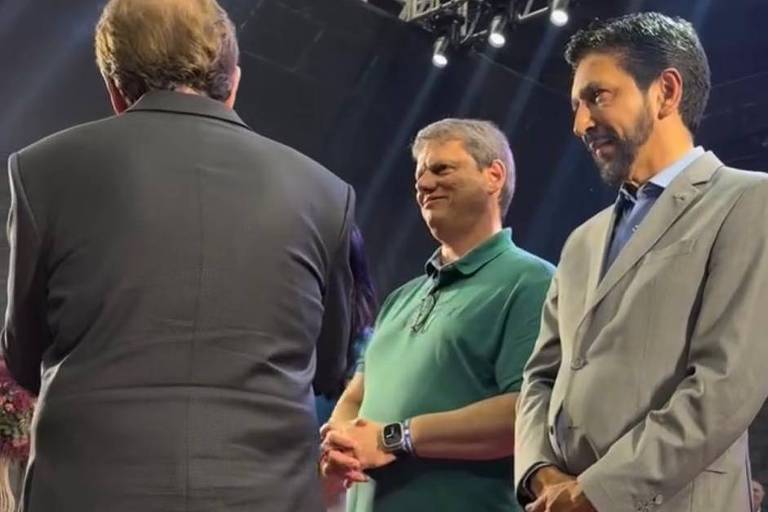 A imagem mostra um grupo de homens em um evento, com foco em três deles. O homem à esquerda está de costas, vestindo um terno escuro. O homem ao centro, com uma camiseta verde, está sorrindo. À direita, um homem com um terno claro e barba está em uma pose pensativa. O fundo é um palco com iluminação e uma decoração floral.