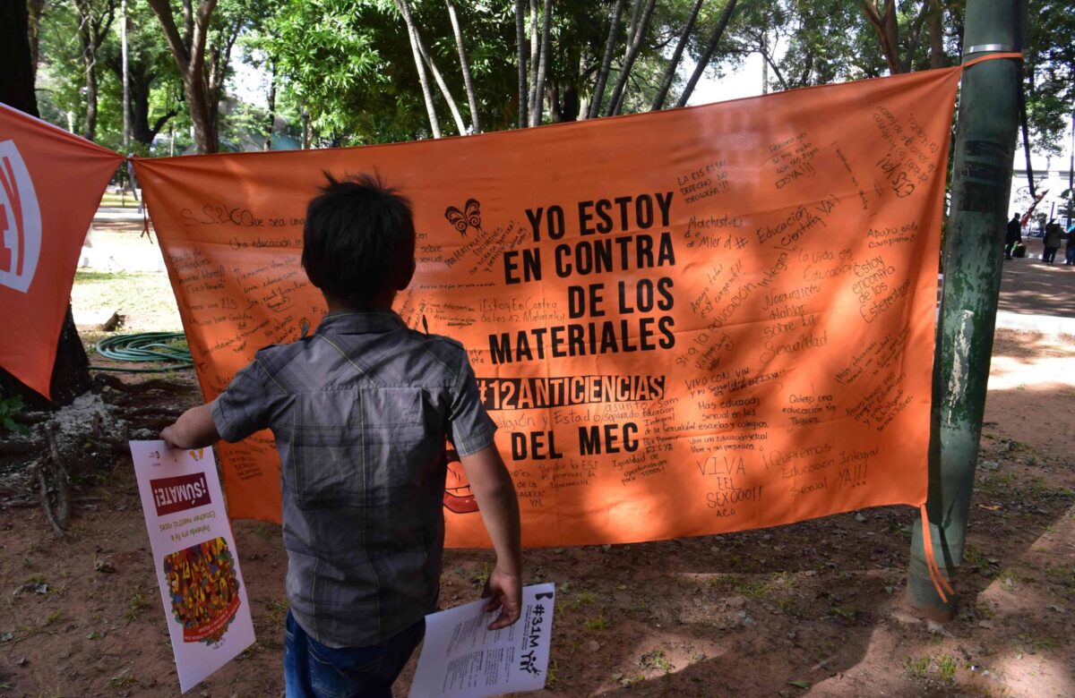 A imagem mostra uma criança de costas, segurando panfletos, em frente a um grande banner laranja. O banner contém a frase