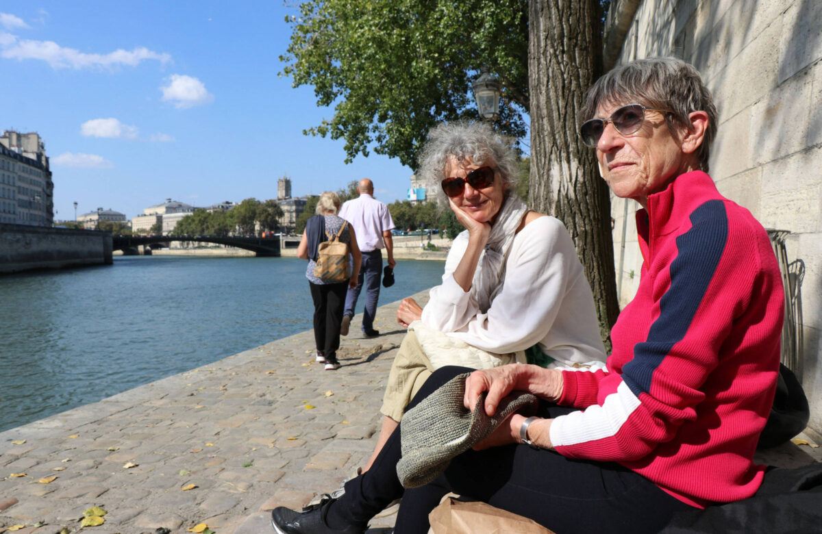 A imagem mostra duas mulheres sentadas em um banco à beira de um rio em Paris. Uma mulher tem cabelo grisalho e usa óculos escuros, enquanto a outra tem cabelo curto e usa uma jaqueta vermelha. Ao fundo, há pessoas caminhando ao longo da margem do rio, com árvores e um edifício visível na distância sob um céu azul com algumas nuvens.