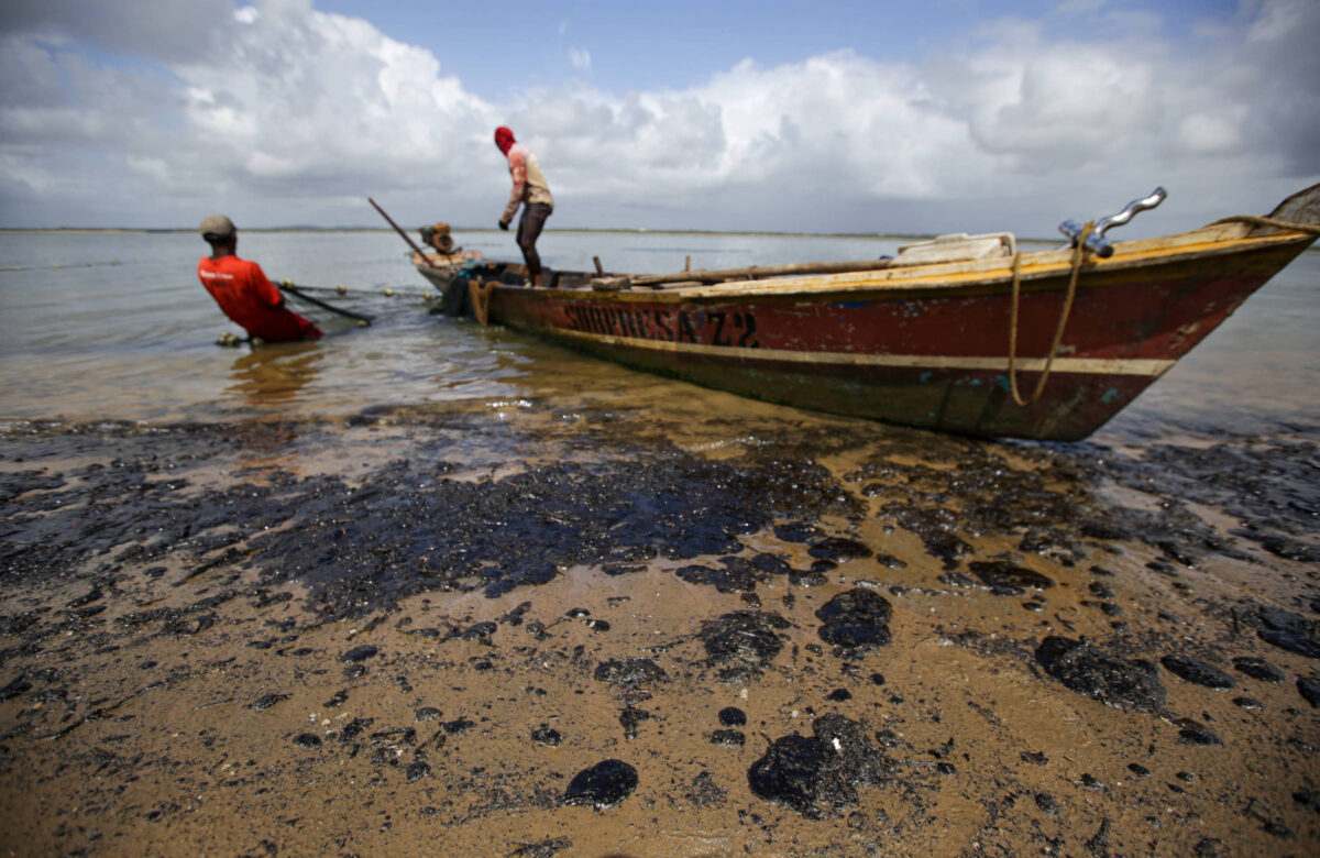 A imagem mostra uma cena em uma praia onde há um barco de madeira parcialmente na água e parcialmente na areia. Duas pessoas estão próximas ao barco: uma pessoa, à esquerda, está na água, inclinada para trás, e a outra, à direita, está em pé sobre o barco. A areia da praia está coberta com manchas escuras de óleo, sugerindo um derramamento de petróleo. O céu ao fundo está parcialmente nublado, com nuvens brancas. A imagem passa uma sensação de impacto ambiental, provavelmente relacionado à contaminação por óleo nas águas e na costa.