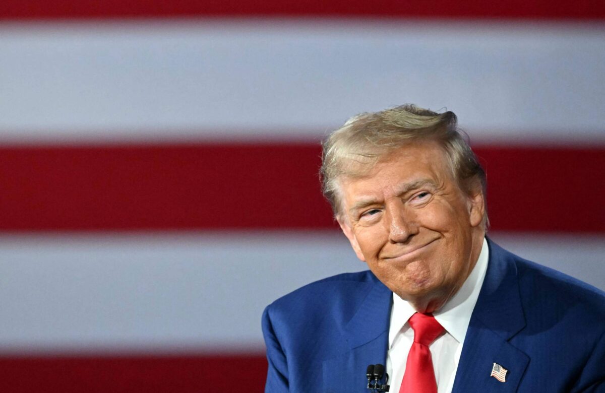 Um homem sorrindo, vestido com um terno azul e uma gravata vermelha, está sentado em frente a uma bandeira com listras vermelhas e brancas. O fundo é predominantemente vermelho com listras brancas