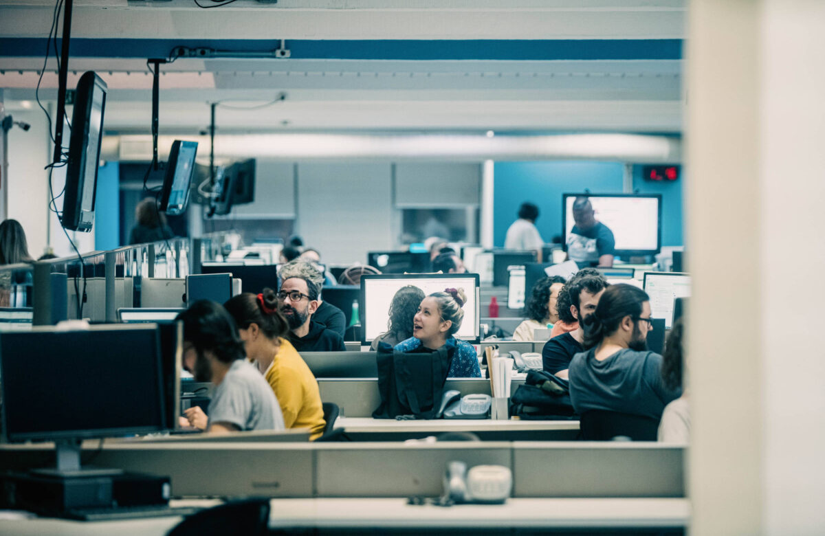 A imagem mostra a Redação da Folha, um ambiente de escritório com várias pessoas trabalhando. Há computadores em cada mesa e algumas pessoas estão interagindo entre si. O espaço é iluminado e parece ser um ambiente colaborativo.