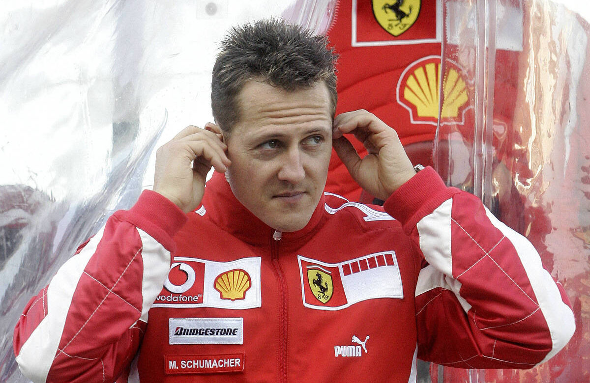 Imagem de um piloto de Fórmula 1 vestindo um uniforme vermelho da Ferrari. O uniforme possui logotipos da Vodafone, Shell, Bridgestone, Puma e Ferrari. O piloto está ajustando algo em seus ouvidos e tem uma expressão concentrada. Ao fundo, há uma imagem ampliada do logotipo da Ferrari e da Shell.