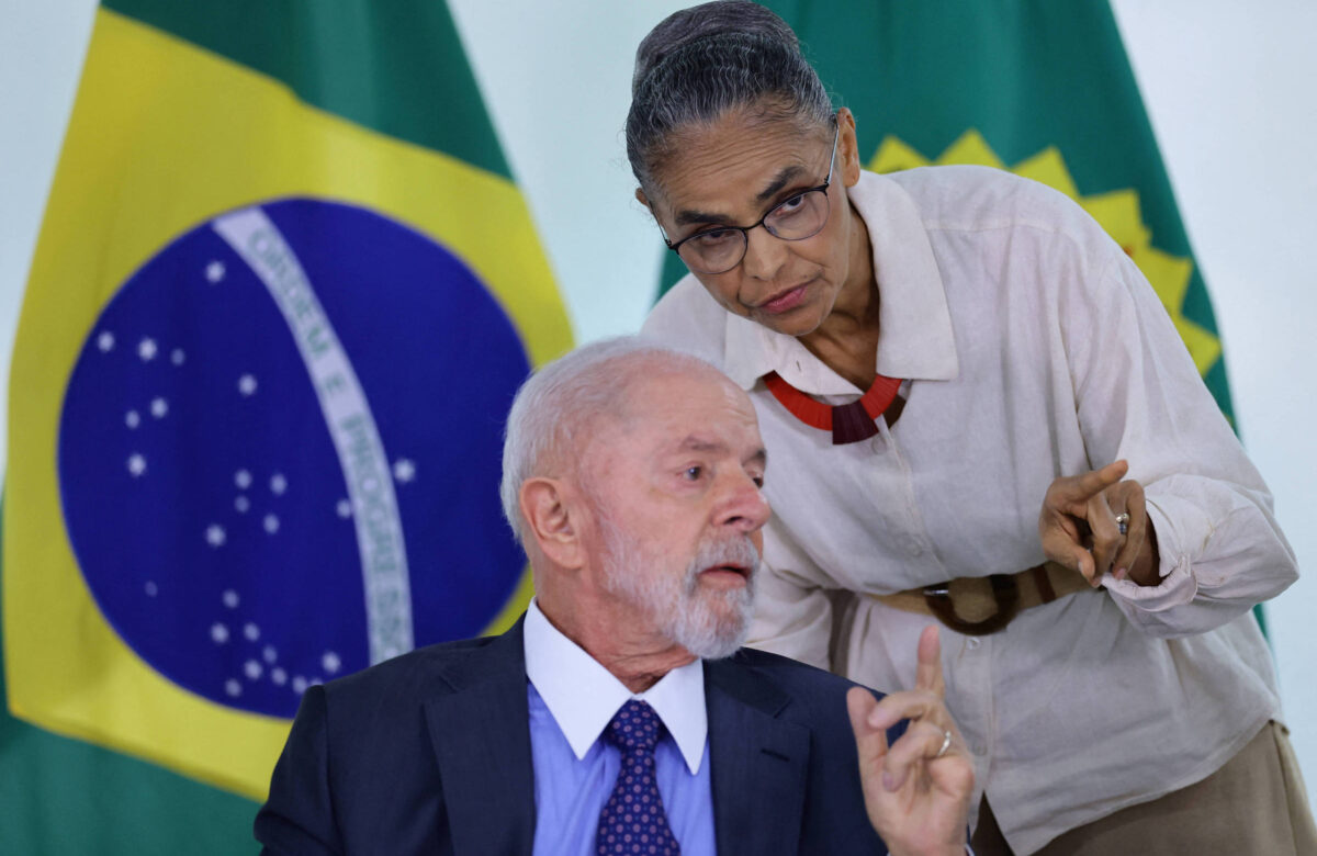 A imagem mostra Lula e Marina Silva em uma conversa. À esquerda, Lula, um homem de cabelo grisalho e barba, vestindo um terno escuro e uma camisa clara, está sentado e gesticulando com a mão. À direita, Marina, com cabelo preso e óculos, vestindo uma blusa clara e um colar, está inclinada para frente, parecendo explicar algo ao presidente. Ao fundo, estão visíveis as bandeiras do Brasil.