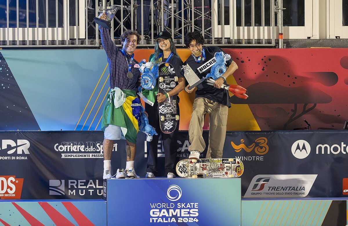 Três skatistas estão em um pódio durante a premiação dos Jogos Mundiais de Skate na Itália. O primeiro skatista, à esquerda, segura um troféu e está vestido com uma camiseta e calças curtas, com uma bandeira do Brasil amarrada na cintura. A skatista do meio, com cabelo azul, segura um skate e está vestida com uma camiseta preta e calças. O terceiro skatista, à direita, segura um troféu e um skate, vestindo uma camiseta preta e calças claras. Ao fundo, há uma decoração colorida e o logotipo dos Jogos Mundiais de Skate 2024