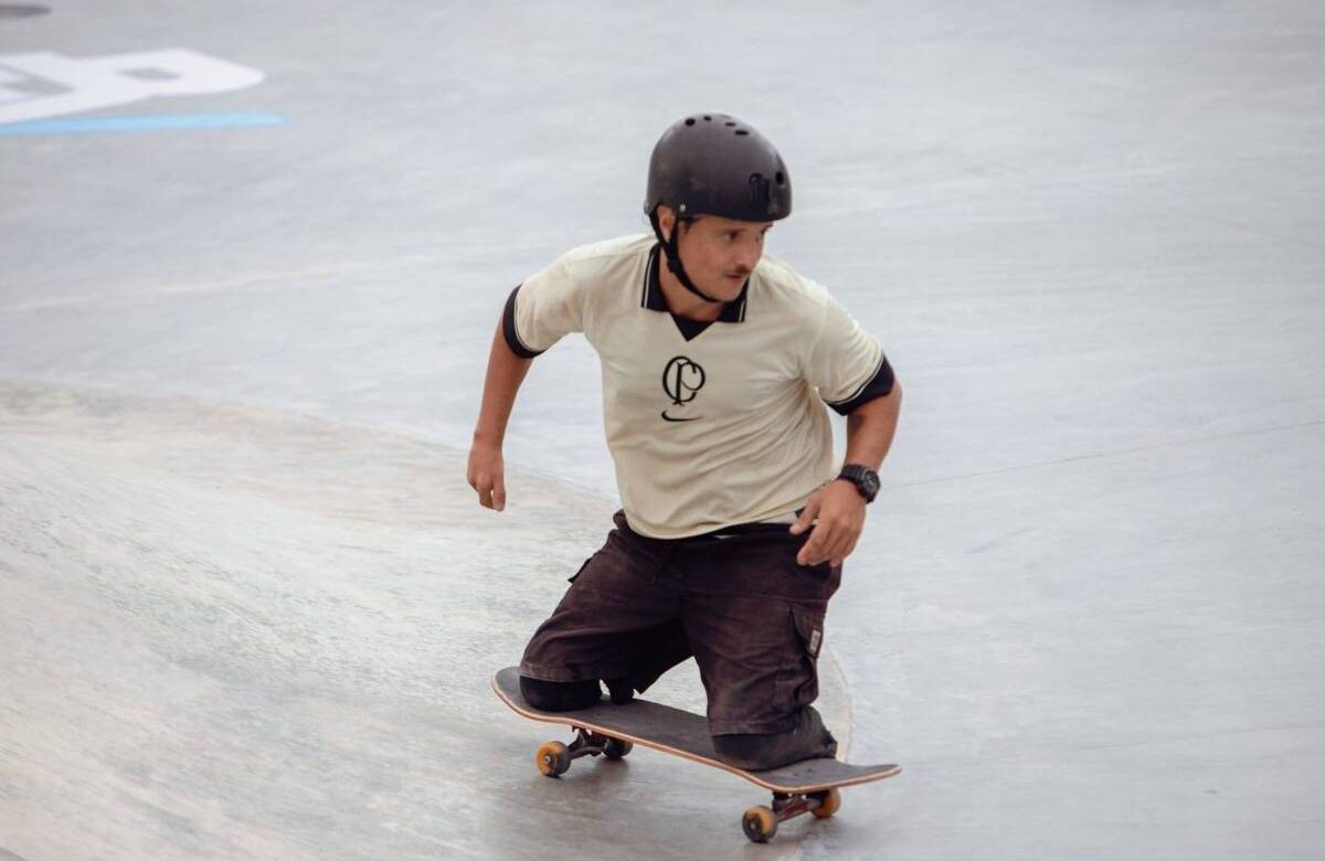 Um skatista está se movendo em uma pista de skate. Ele usa um capacete preto e uma camiseta clara com um logotipo. O skatista está em uma posição inclinada, com um dos joelhos apoiado no skate e o outro pé no chão. O ambiente é uma pista de skate com superfície de concreto.