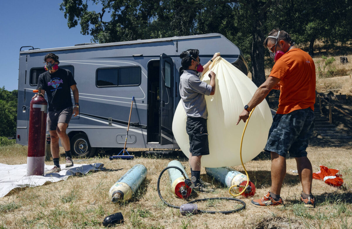 A imagem mostra três pessoas em um campo, próximas a um veículo recreativo. Uma pessoa está segurando um cilindro de gás, enquanto outra está inflando um balão grande e amarelo claro. A terceira pessoa está apontando para o balão. Todos estão usando máscaras de proteção. No chão, há cilindros de gás e uma lona branca.