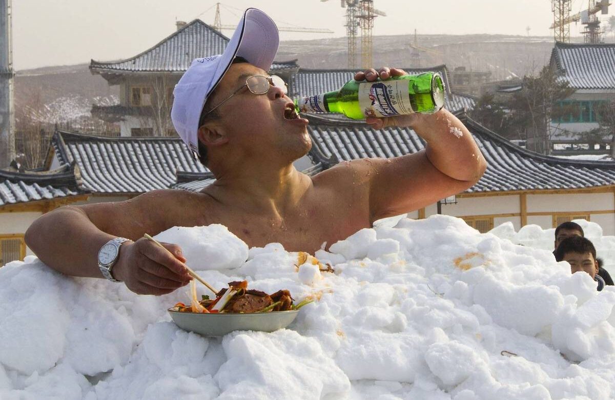 Homem sem camisa, enterrado num monte de neve, bebe cerveja