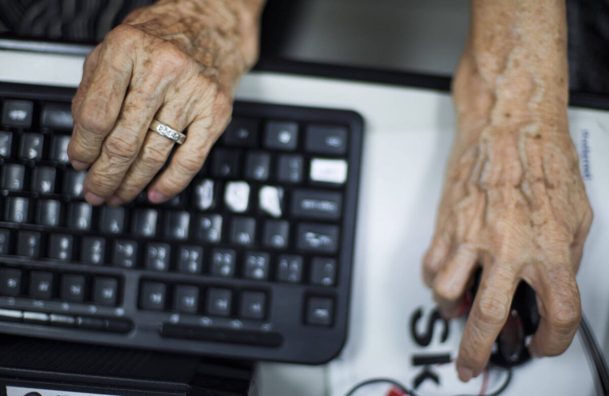 A imagem mostra as mãos de uma pessoa idosa sobre um teclado preto e um mouse. As mãos são enrugadas e uma delas usa um anel. O fundo é desfocado, mas é possível ver uma superfície clara sob o teclado.