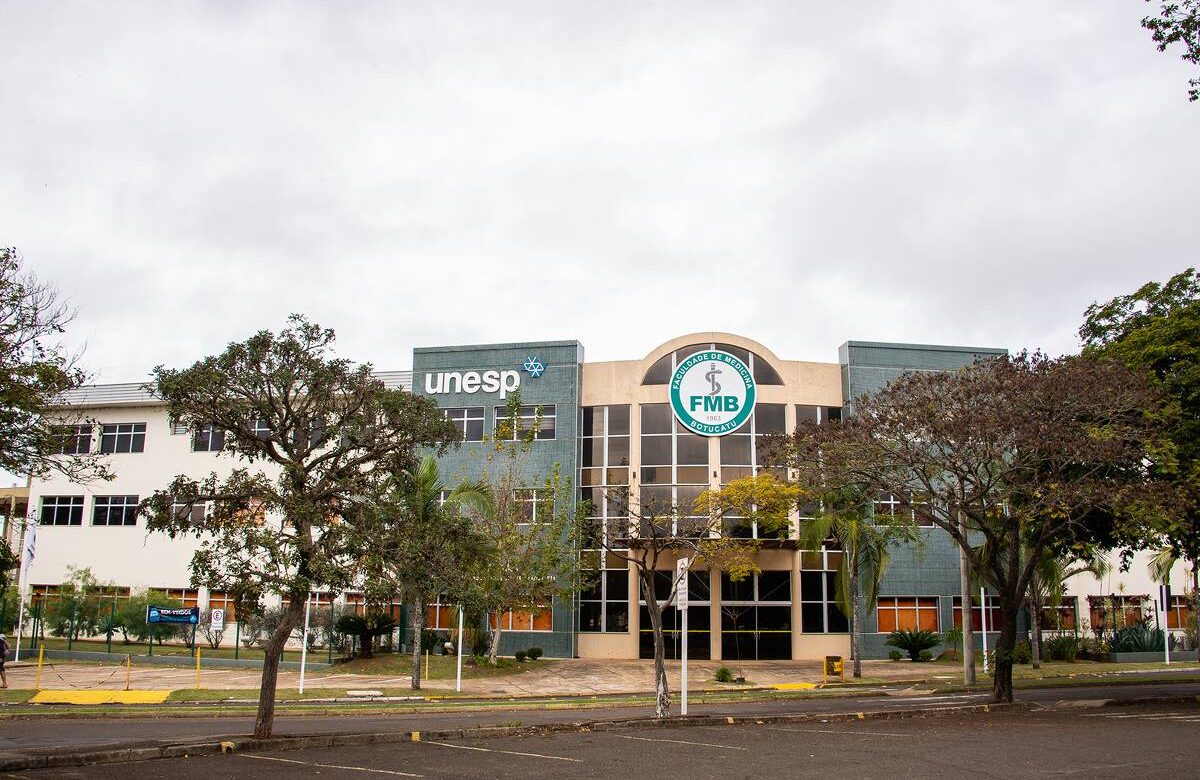 Imagem da fachada de um prédio da UNESP, com o logotipo da universidade visível. O edifício é de dois andares, com janelas grandes e uma entrada central. À frente, há árvores e um espaço pavimentado. O céu está nublado.