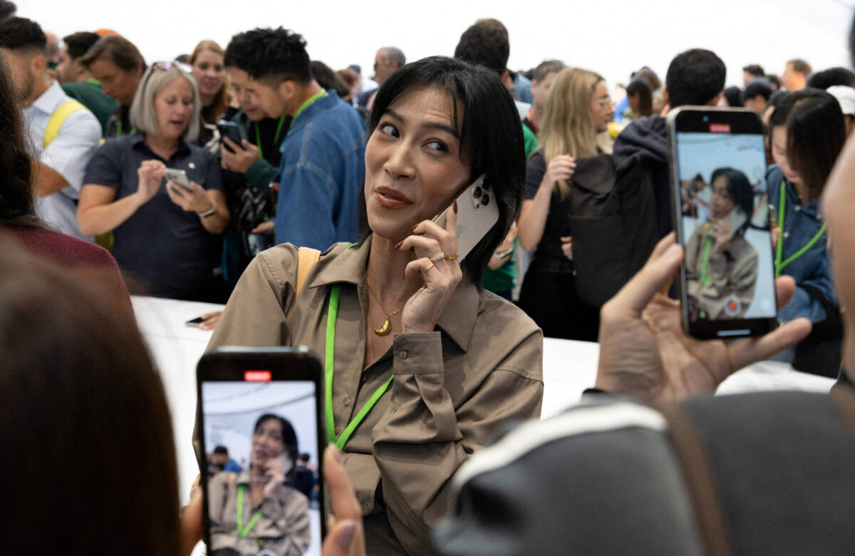 A imagem mostra uma mulher em primeiro plano, falando ao telefone e olhando para a câmera. Ao fundo, há um grupo de pessoas, algumas delas também segurando smartphones. A mulher está vestindo uma camisa bege e tem cabelo escuro. O ambiente parece ser um evento ou conferência, com uma grande quantidade de pessoas ao redor.