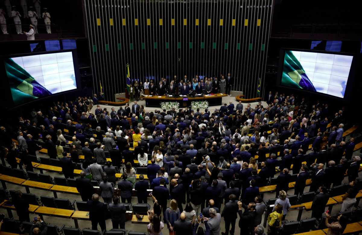 A imagem mostra uma sessão plenária na Câmara dos Deputados do Brasil. Há um grande número de deputados em pé, em um ambiente com cadeiras dispostas em fileiras. No fundo, há telas de projeção exibindo informações. O ambiente é bem iluminado e decorado com as cores da bandeira brasileira.
