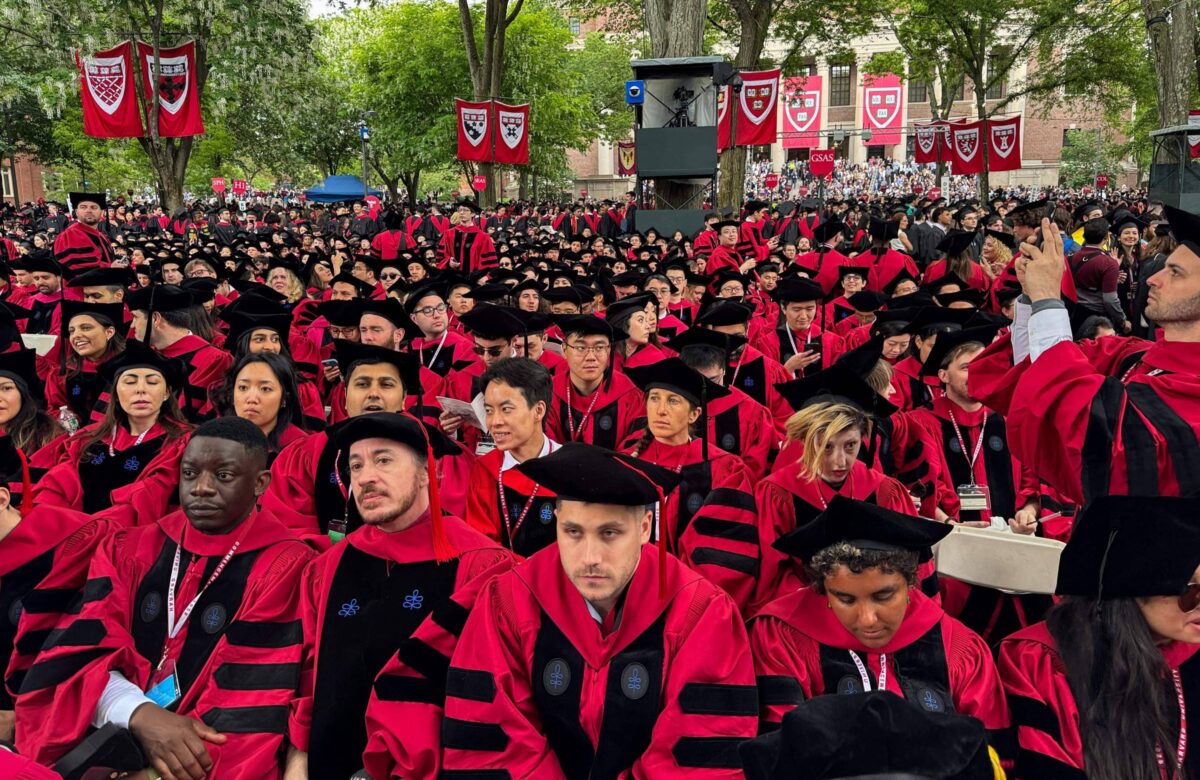 A imagem mostra uma grande multidão de graduados sentados em um evento de formatura. Eles estão vestidos com togas e chapéus de formatura, predominantemente em vermelho e preto. Ao fundo, há bandeiras de uma instituição de ensino, possivelmente Harvard, e árvores ao redor. A atmosfera é de celebração e solenidade, com muitos graduados olhando para frente e alguns interagindo entre si.