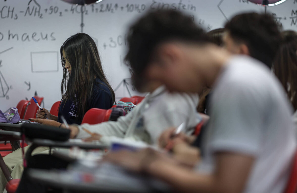 A imagem mostra um grupo de estudantes em uma sala de aula. No primeiro plano, uma estudante com cabelo longo e escuro está concentrada, escrevendo em um caderno. Ao fundo, outros estudantes estão visíveis, alguns com a cabeça baixa, também escrevendo. As mesas são de cor escura e as cadeiras são vermelhas. As paredes da sala têm anotações e fórmulas científicas escritas, sugerindo um ambiente acadêmico.
