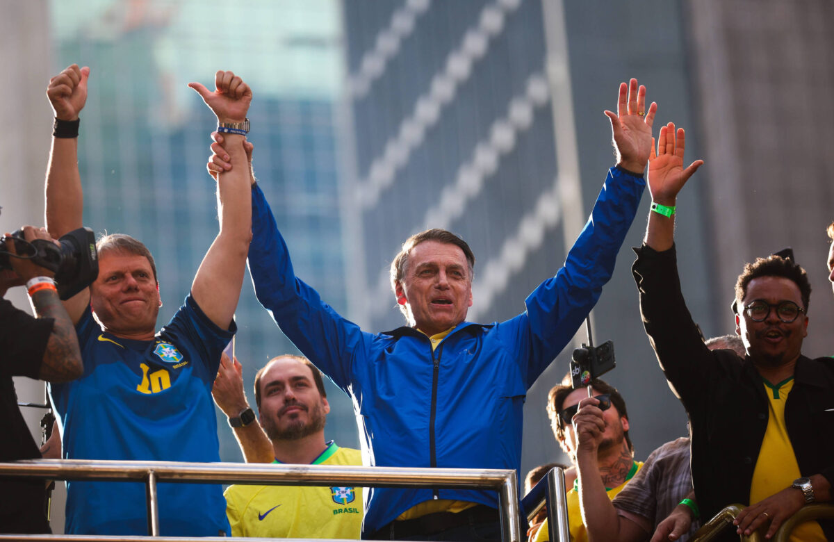 Um grupo de homens está em um palco, com um deles levantando as mãos em sinal de vitória. Eles estão vestidos com camisetas da seleção brasileira de futebol. O homem no centro, que usa uma jaqueta azul, parece estar se dirigindo a uma multidão. Ao fundo, há edifícios altos e uma atmosfera de celebração.