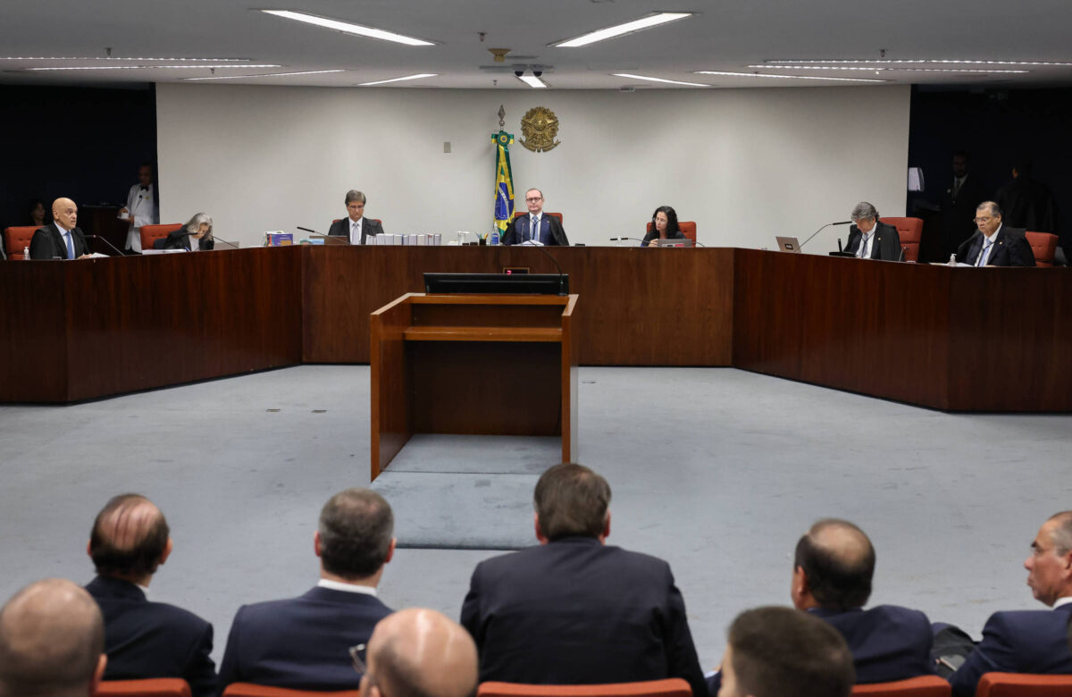 A imagem mostra uma sessão em um plenário, com um grupo de pessoas sentadas em cadeiras dispostas em um formato semicircular. No centro, há uma mesa retangular vazia. Ao fundo, há uma bandeira do Brasil e o brasão nacional. Os participantes estão vestidos formalmente, e a sala possui iluminação artificial.