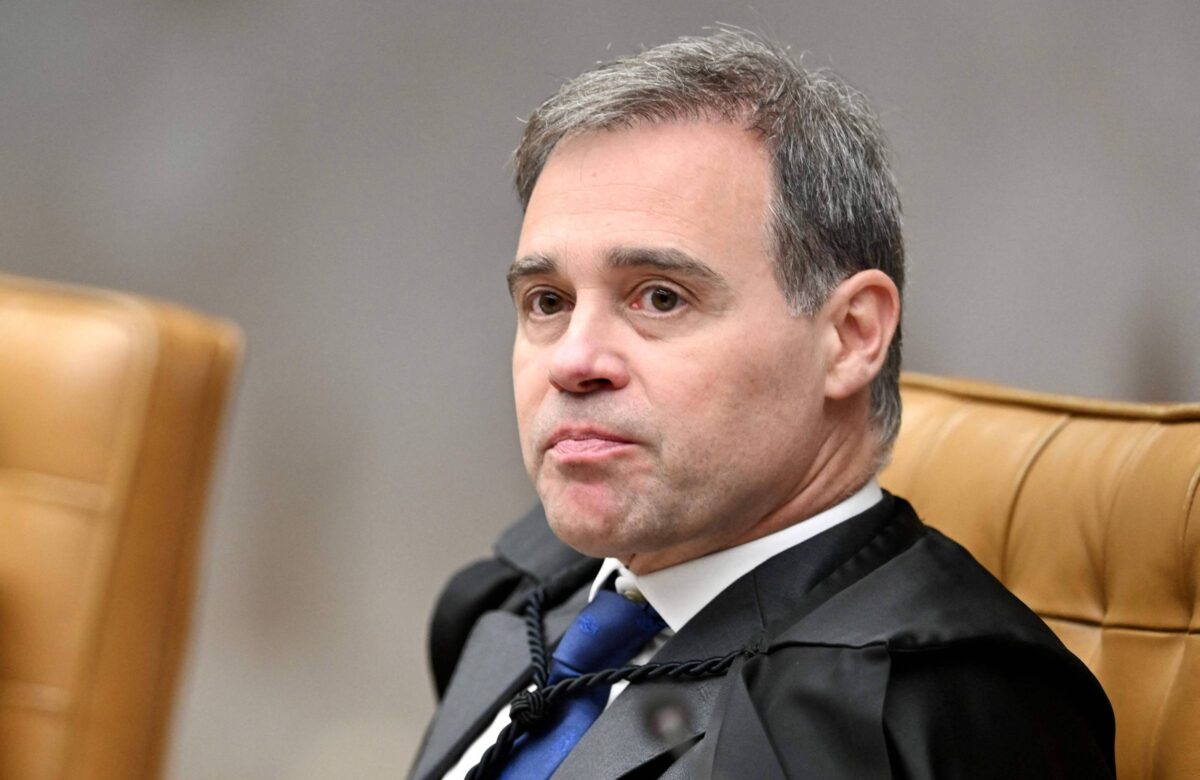 Um homem com cabelo curto e grisalho está sentado em uma cadeira de tribunal. Ele usa uma toga preta e uma gravata azul. O fundo é desfocado, mas parece ser um ambiente de tribunal, com cadeiras de couro marrom ao redor. O homem tem uma expressão séria e está olhando para o lado.