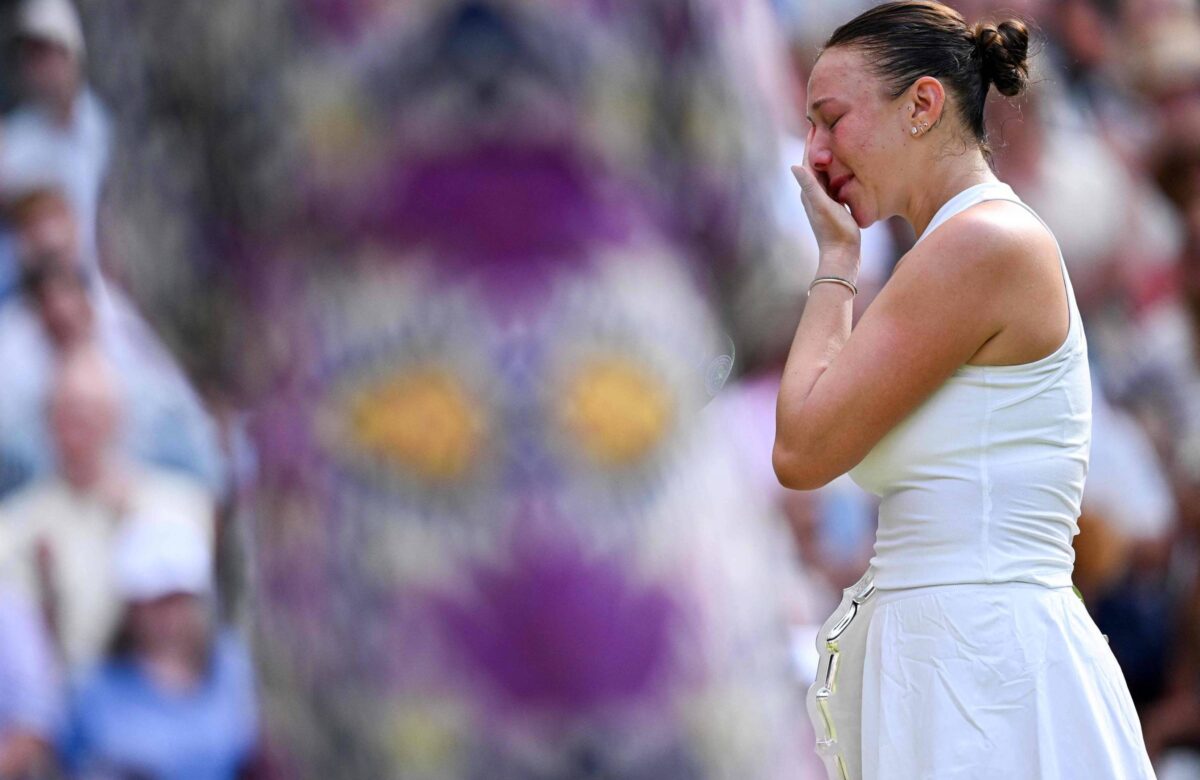 Uma jogadora de tênis está em pé, vestindo um vestido branco, com uma expressão de emoção no rosto. Ela está limpando uma lágrima com a mão, enquanto observa o público ao fundo. O cenário é de um torneio de tênis, com espectadores visíveis, mas desfocados, ao fundo. A atmosfera parece ser de tensão e emoção, possivelmente após uma partida importante.