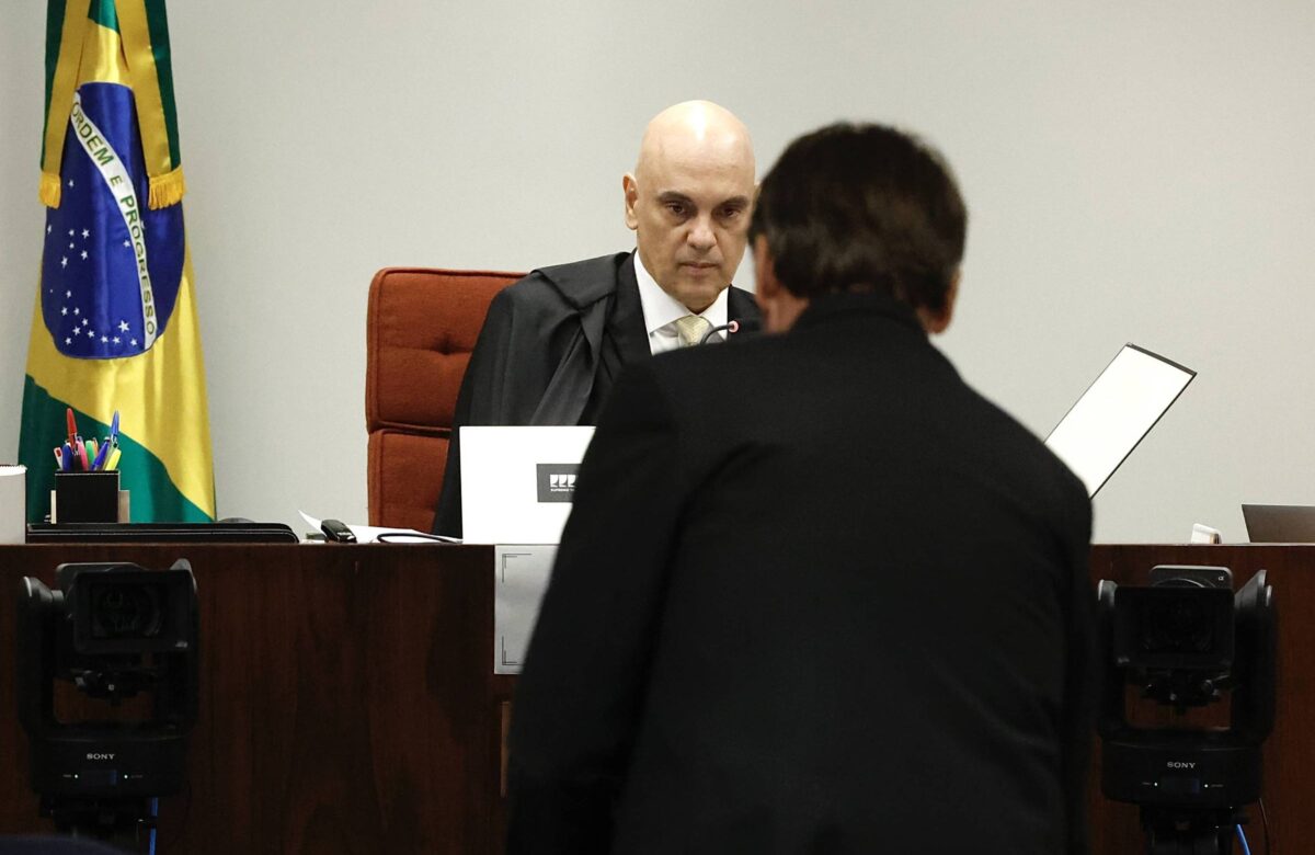 A imagem mostra um juiz com cabelo calvo e vestindo uma toga, sentado em uma mesa de tribunal. Ele está olhando para um homem de costas, que está de pé em frente a ele. Ao fundo, há uma bandeira do Brasil e equipamentos de gravação. O ambiente é de um tribunal, com uma atmosfera formal.