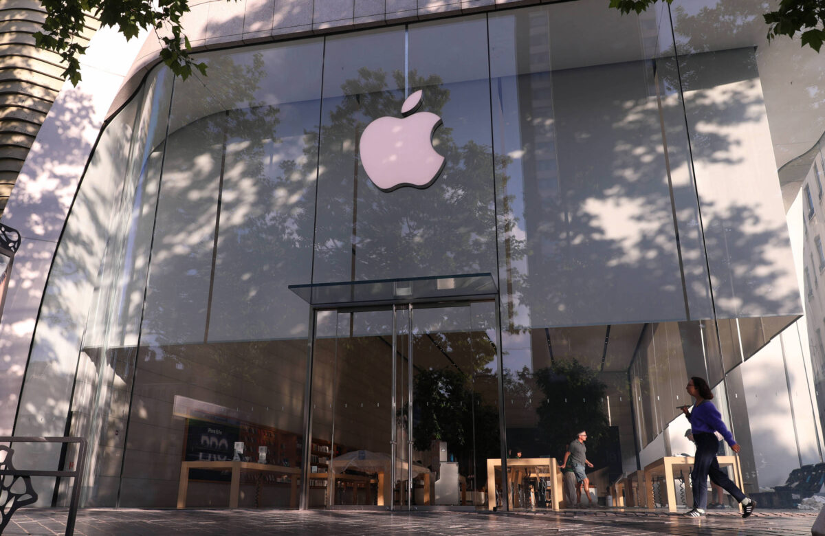 A imagem mostra a fachada de uma loja da Apple, com grandes janelas de vidro e o logotipo da Apple em destaque na parte superior. Algumas pessoas estão caminhando em frente à loja, e há mesas de exposição visíveis dentro do estabelecimento.