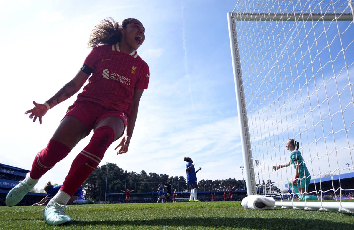 Uma jogadora de futebol, vestindo um uniforme vermelho do Liverpool, está comemorando um gol. Ela está em primeiro plano, com uma expressão de alegria e os braços levantados. Ao fundo, outras jogadoras estão em campo, e uma goleira é visível. O céu está claro e ensolarado, e o gramado parece bem cuidado.