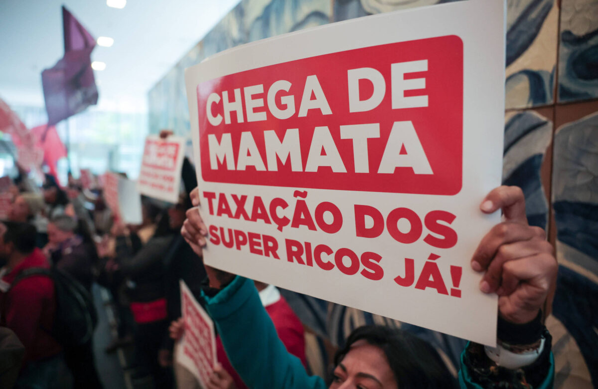 A imagem mostra um grupo de pessoas participando de um protesto. Em primeiro plano, uma mulher segura um cartaz com as inscrições