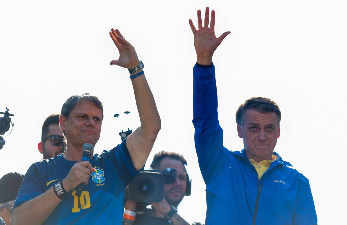 Duas pessoas estão levantando a mão em um evento público. À esquerda, um homem com uma camiseta azul e o número 10, e à direita, um homem com uma jaqueta azul e uma camisa amarela. Ao fundo, há várias pessoas e câmeras registrando o momento.