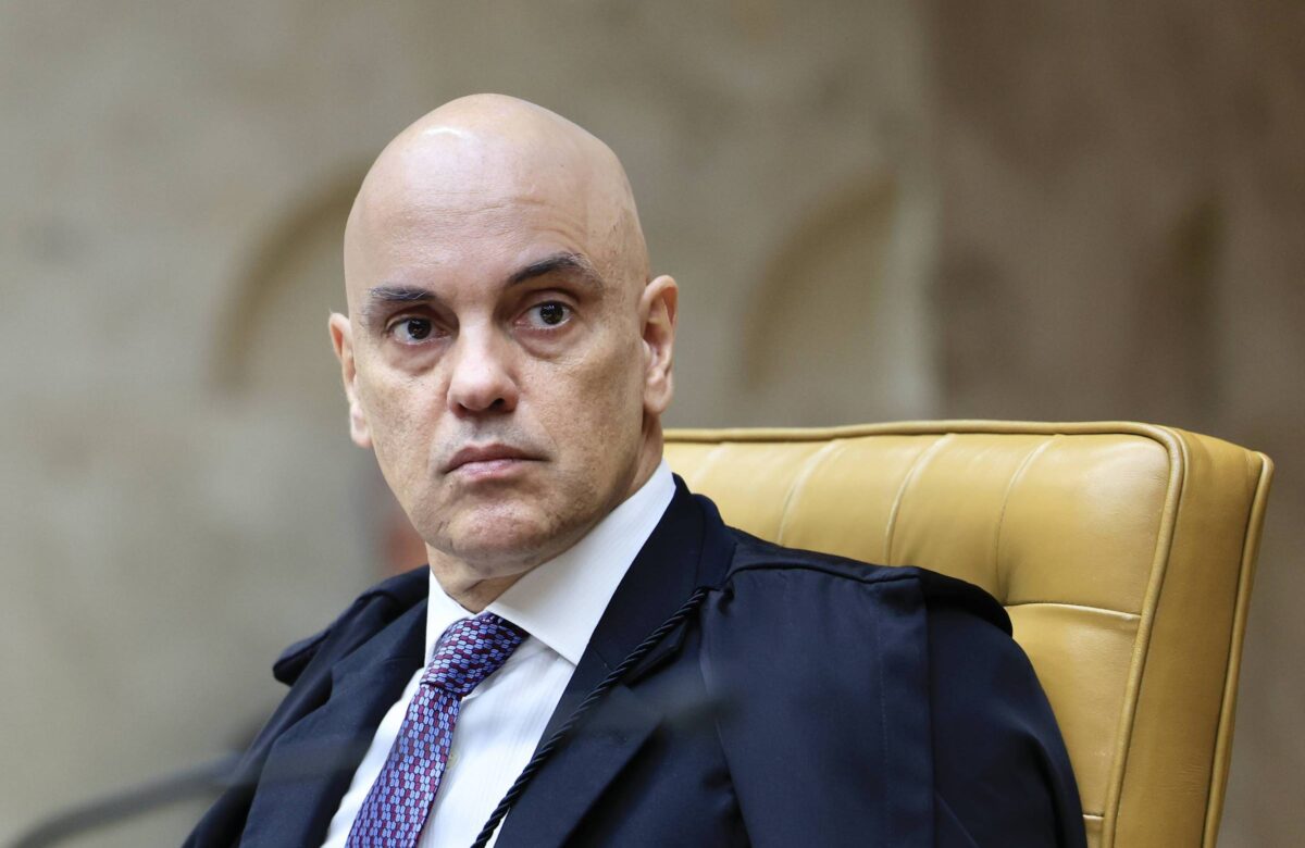 Um homem calvo, com pele clara, está sentado em uma cadeira amarela. Ele usa um terno escuro e uma gravata roxa. O fundo é desfocado, mas parece ser um ambiente formal, possivelmente um tribunal. O homem tem uma expressão séria e está olhando para frente.