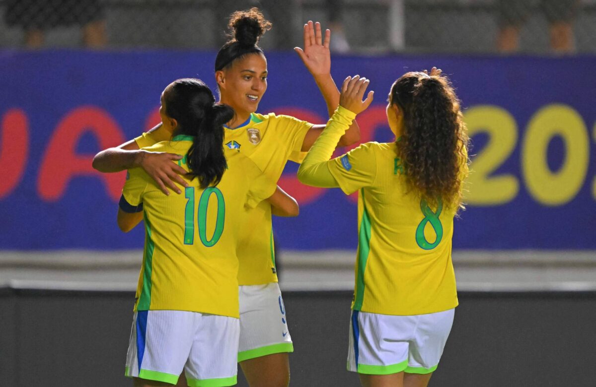Três jogadoras de futebol feminino estão se abraçando e comemorando em um campo. Elas vestem uniformes amarelos com detalhes em verde e azul. A jogadora à esquerda, com o número 10, está abraçando a jogadora do meio, que tem o cabelo preso. A jogadora à direita, com o número 8, levanta a mão em celebração. Ao fundo, há uma faixa colorida com a inscrição