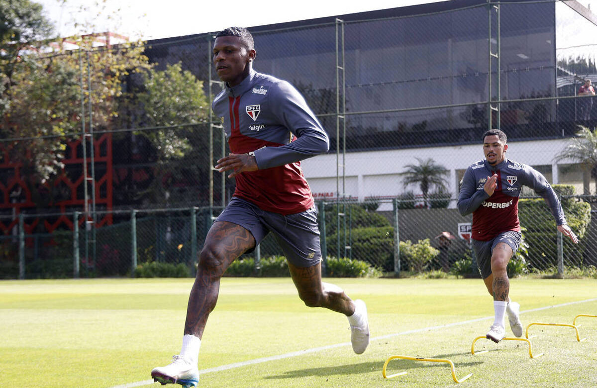 Dois jogadores de futebol estão correndo em um campo de treinamento. O primeiro jogador, à frente, tem tatuagens visíveis e está usando uma camisa de treino vermelha e cinza. O segundo jogador, atrás, também está vestido com uma camisa semelhante e parece estar se esforçando para alcançar o primeiro. Ao fundo, há uma estrutura de vidro e algumas árvores.
