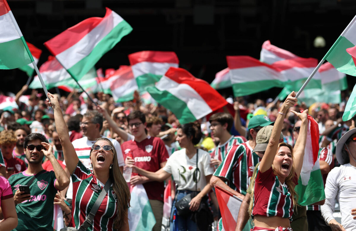Uma multidão de torcedores celebra em um estádio, segurando bandeiras nas cores verde, branco e vermelho. As pessoas estão vestidas com camisas de futebol, algumas com listras nas mesmas cores. O ambiente é festivo, com muitos torcedores sorrindo e levantando os braços em comemoração.