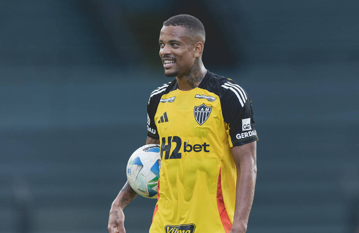 Um jogador de futebol está sorrindo enquanto segura uma bola. Ele veste uma camisa de goleiro predominantemente amarela com detalhes em preto e laranja, e o logotipo do time