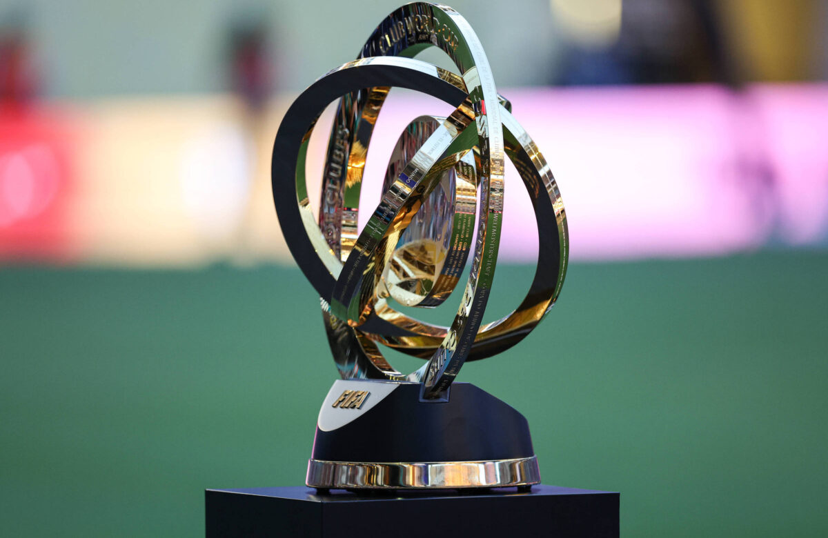 A imagem mostra um troféu de futebol com um design moderno, composto por anéis entrelaçados em dourado e prateado, posicionado sobre uma base preta. O fundo é desfocado, com luzes coloridas ao fundo, sugerindo um ambiente de evento esportivo.