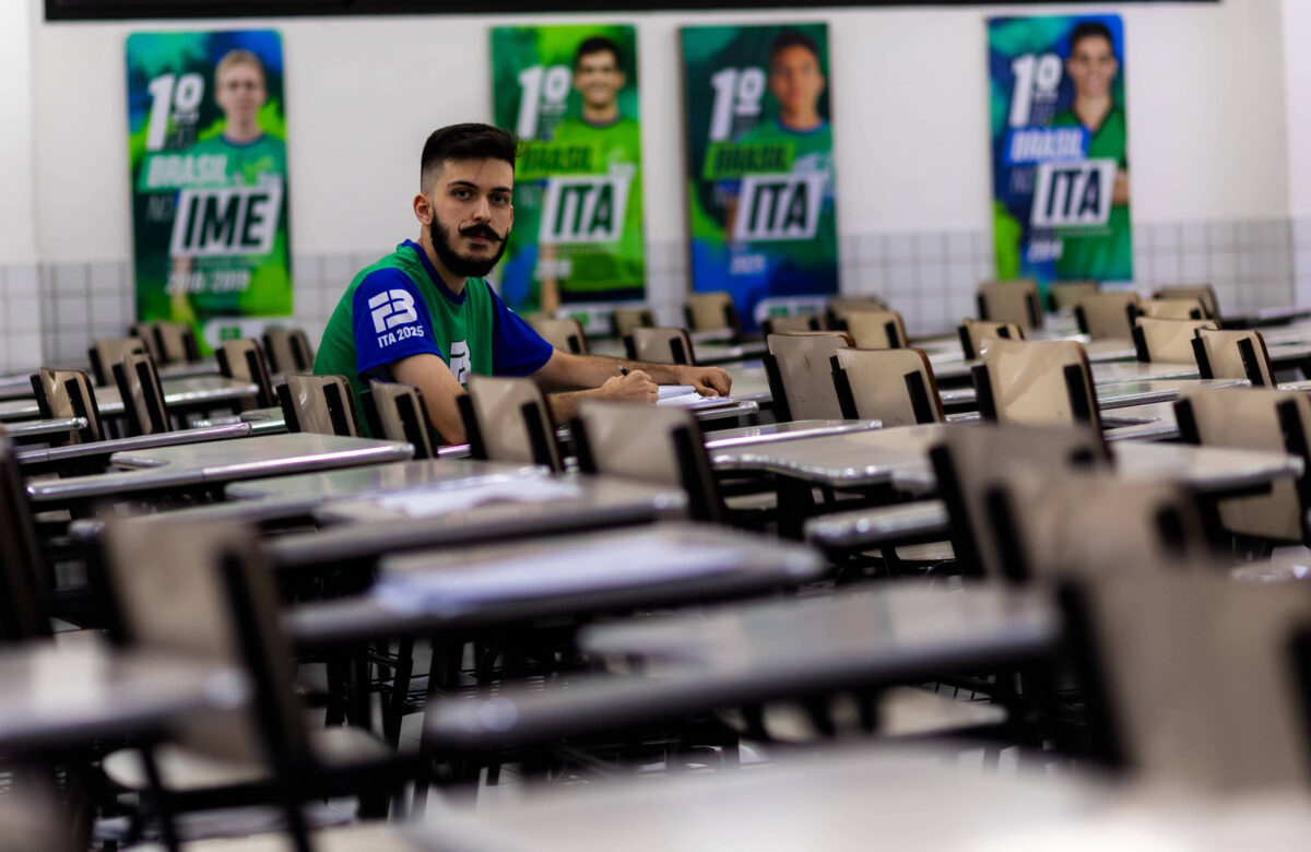 A imagem mostra um jovem sentado em uma mesa em um refeitório vazio. Ele está usando uma camiseta azul e parece estar escrevendo em um caderno. Ao fundo, há cartazes com os números 1 e 2, que parecem se referir a competições ou eventos, e estão relacionados a