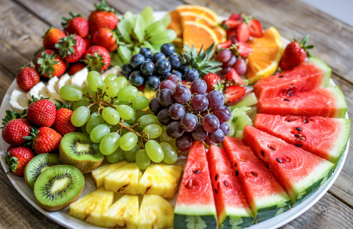 Um prato redondo repleto de diversas frutas frescas. Há morangos, uvas verdes e roxas, fatias de melancia, abacaxi, kiwi, laranjas e uvas. As frutas estão dispostas de forma colorida e atraente, com uma variedade de texturas e cores.